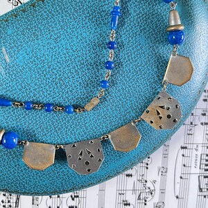 May include: A vintage necklace with a silver chain and blue beads. The necklace features silver pendants with cut-out designs. The jewelry is displayed on a textured blue surface, with sheet music in the background.