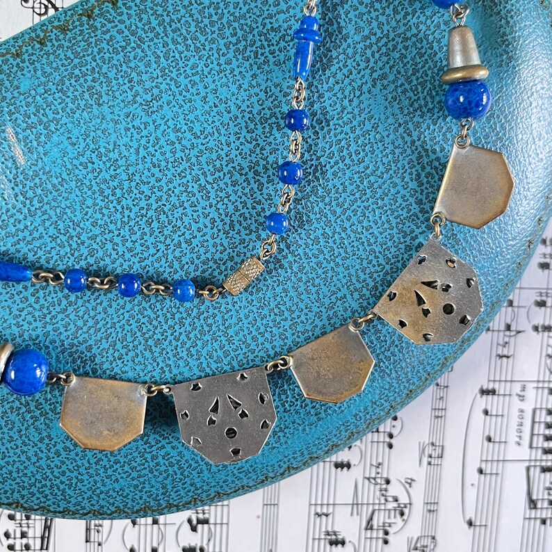 May include: A vintage necklace with a silver chain and blue beads. The necklace features silver pendants with cut-out designs. The jewelry is displayed on a textured blue surface, with sheet music in the background.
