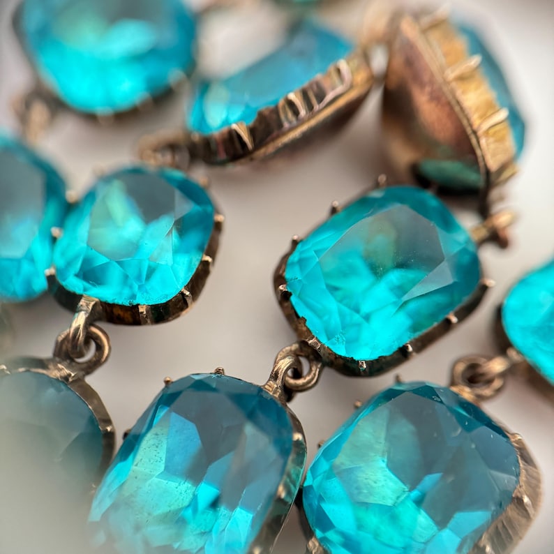 May include: A necklace featuring multiple faceted, rectangular bright turquoise stones set in metallic frames. Each stone is connected by small metal links, creating a cascading arrangement of vibrant blue gems with visible facets reflecting light.