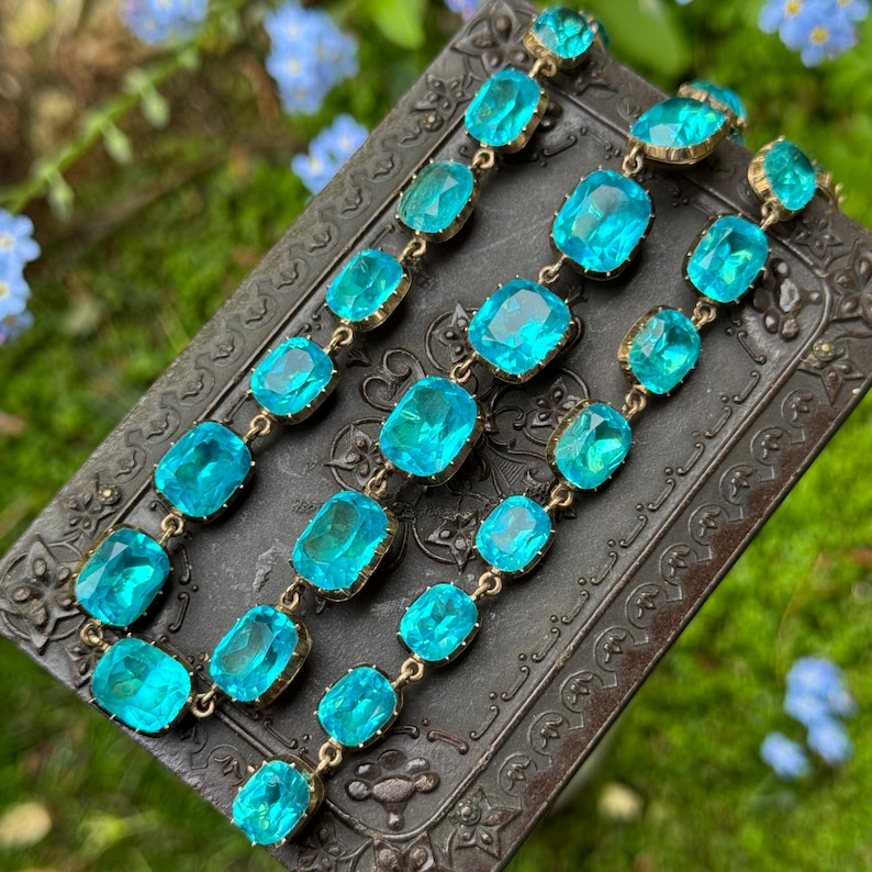 May include: A bracelet featuring multiple rectangular, faceted, bright turquoise-blue glass stones set in gold-toned metal frames. The stones are linked together in a continuous chain, showcasing a vibrant, translucent color and reflective surfaces.