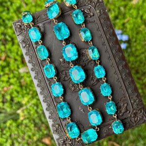May include: A long necklace featuring three parallel strands of faceted, rectangular, bright turquoise blue glass stones set in gold-toned metal frames. The stones are arranged in a repeating pattern, creating a vibrant, shimmering effect.