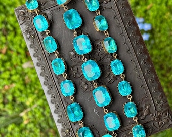 May include: A long necklace featuring three parallel strands of faceted, rectangular, bright turquoise blue glass stones set in gold-toned metal frames. The stones are arranged in a repeating pattern, creating a vibrant, shimmering effect.
