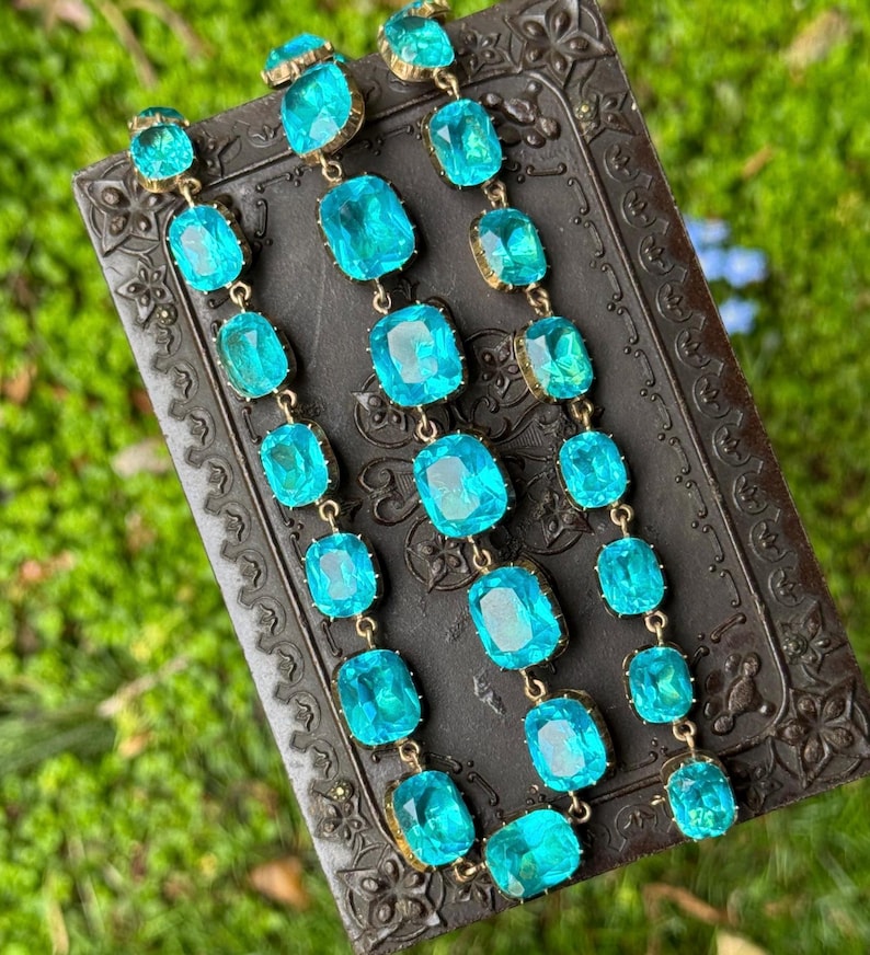 May include: A long necklace featuring three parallel strands of faceted, rectangular, bright turquoise blue glass stones set in gold-toned metal frames. The stones are arranged in a repeating pattern, creating a vibrant, shimmering effect.