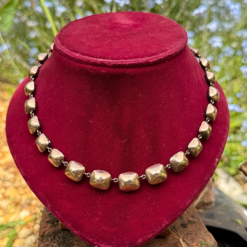 May include: A necklace featuring a series of polished, metallic gold-toned square beads linked together in a continuous circular chain. Each bead has a smooth, slightly rounded surface that reflects light, creating a uniform and repeating pattern.