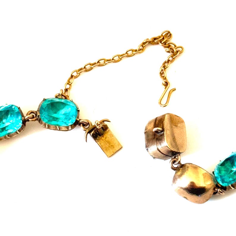 May include: A gold-toned necklace featuring a series of oval-shaped, vibrant turquoise-blue faceted stones set in metal bezels. The piece includes a metal chain extension with a hook clasp and a rectangular box-style clasp mechanism for closure.