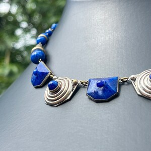 May include: A blue necklace with silver-colored metal accents. The necklace features geometric shapes and round beads. The focal point includes a large, dark blue, faceted stone. The necklace is displayed on a black mannequin.