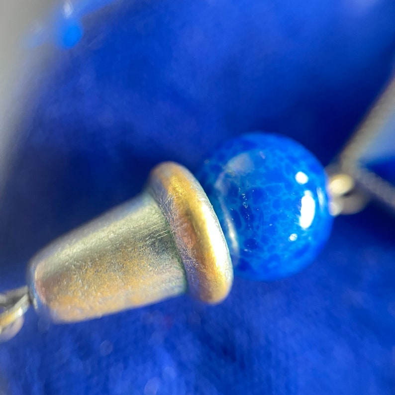 May include: Close-up of a silver-colored metal bead cap with a gold-colored rim, connected to a blue, textured bead. The background is a deep blue fabric, creating a contrast with the metallic and blue elements. The image highlights the details of the jewelry.