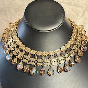 Könnte beinhalten: Eine goldfarbene Choker-Halskette mit mehrlagigem Ketten-Design und baumelnden, regenbogenfarbenen Kristallen in Tropfenform.