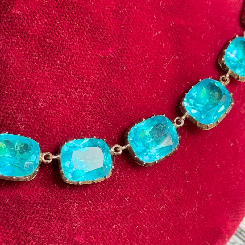 May include: A necklace featuring a series of square-cut, bright turquoise blue stones set in metal frames. Each stone is linked together by small metal connectors, creating a flexible chain of faceted gems that catch the light.