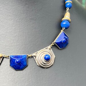 May include: A close-up of a vintage necklace featuring deep blue lapis lazuli stones and silver-toned metal accents. The necklace has geometric and spiral designs, with a mix of round and faceted stones. The background is a dark gray.