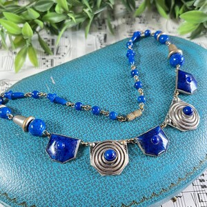 May include: A vintage necklace featuring a combination of blue lapis lazuli beads and geometric silver-toned pendants. The necklace has a central chain of small blue beads and larger blue stone accents. The pendants have a spiral design.