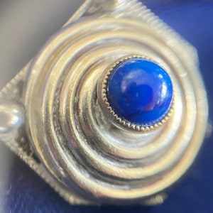 May include: A silver-toned, spiral-shaped jewelry piece with a vibrant blue gemstone at its center. The gemstone is encircled by a gold-colored setting, adding a touch of contrast. The design features a textured, swirling pattern.