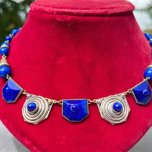 May include: A striking necklace featuring vibrant blue lapis lazuli beads and geometric pendants. The necklace has a silver-toned metal setting with a central spiral design and a series of blue stone accents. The necklace is displayed on a red velvet surface.