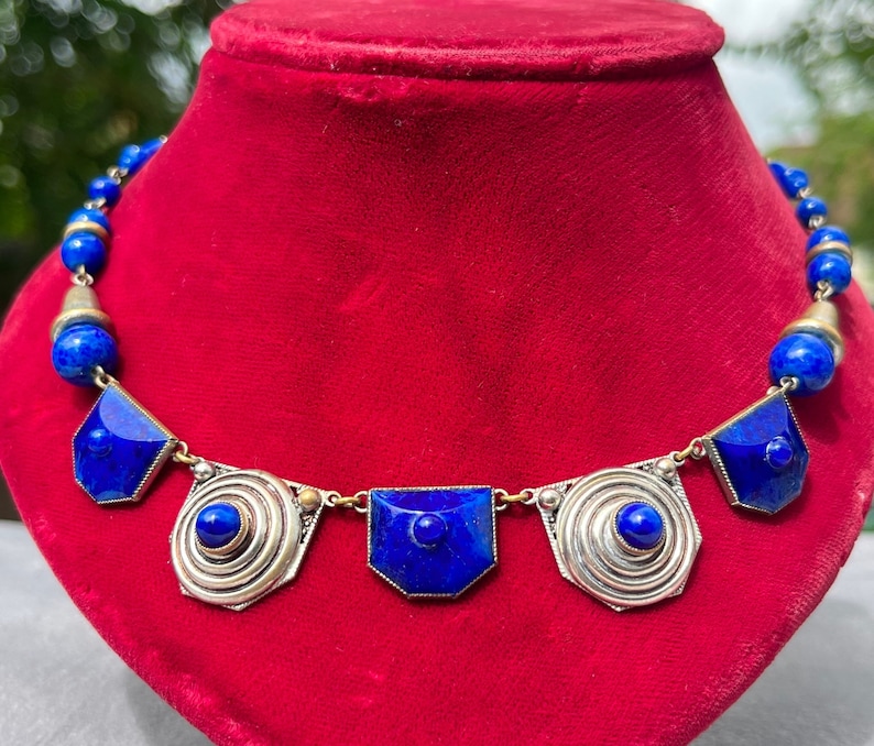 May include: A striking necklace featuring vibrant blue lapis lazuli beads and geometric pendants. The necklace has a silver-toned metal setting with a central spiral design and a series of blue stone accents. The necklace is displayed on a red velvet surface.