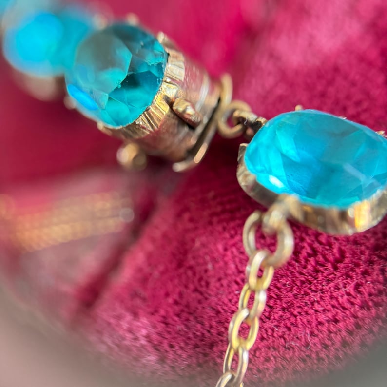 May include: A bracelet featuring a series of faceted, bright turquoise-colored stones set in gold-toned metal prongs. The stones are connected by small, delicate gold-toned metal links, creating a flexible chain structure that catches the light.