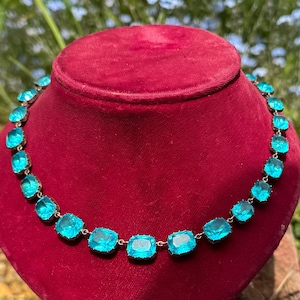 May include: A necklace featuring a single row of square-cut, bright turquoise blue stones set in a silver-toned metal frame. The stones are linked together in a continuous chain pattern, creating a uniform design that encircles the neck display.