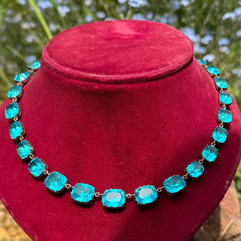 May include: A necklace featuring a single row of square-cut, bright turquoise blue stones set in a silver-toned metal frame. The stones are linked together in a continuous chain pattern, creating a uniform design that encircles the neck display.