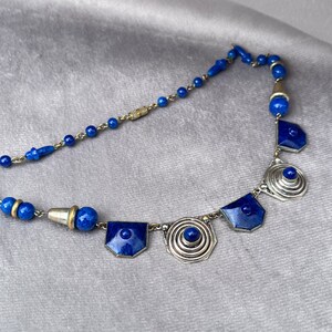 May include: A vintage necklace featuring a combination of blue beads and geometric silver-toned pendants. The necklace has a delicate chain and a variety of shapes, including round beads and shield-shaped pendants. The blue color is a deep, rich shade.