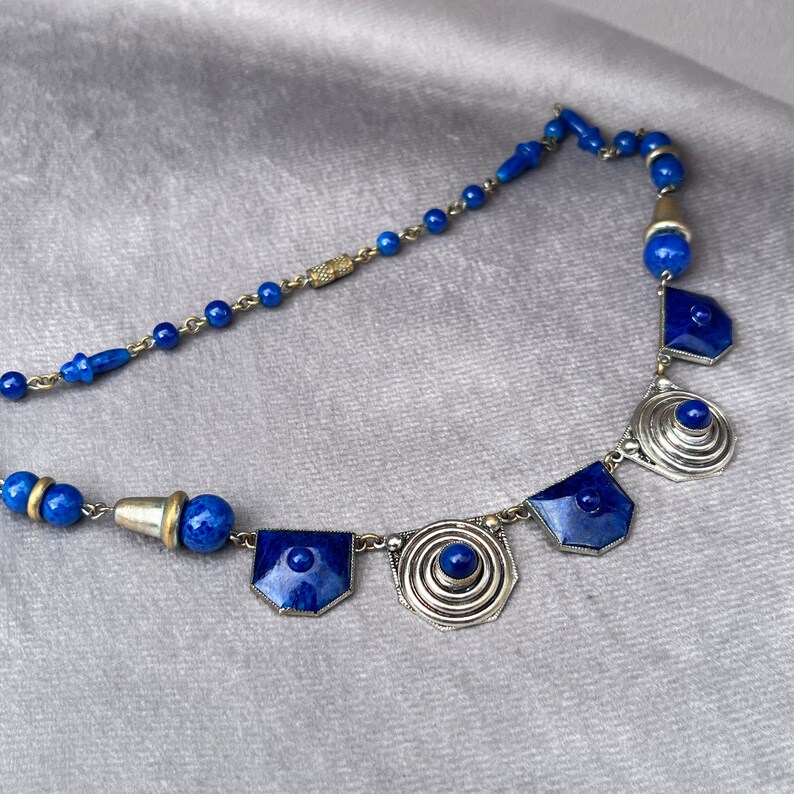 May include: A vintage necklace featuring a combination of blue beads and geometric silver-toned pendants. The necklace has a delicate chain and a variety of shapes, including round beads and shield-shaped pendants. The blue color is a deep, rich shade.