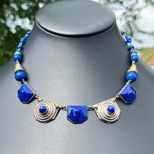 May include: A striking blue necklace with silver-toned metal accents. The necklace features a series of geometric blue stones and round blue beads. The design includes spiral and rectangular shapes, creating a bold and elegant statement piece.