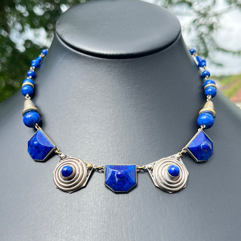 May include: A striking blue necklace with silver-toned metal accents. The necklace features a series of geometric blue stones and round blue beads. The design includes spiral and rectangular shapes, creating a bold and elegant statement piece.