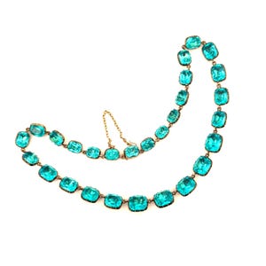 May include: A necklace featuring a series of rectangular, faceted turquoise-colored glass stones set in a metallic frame. The stones are linked together in a continuous chain, with a small metal clasp and a short safety chain attached to the links.