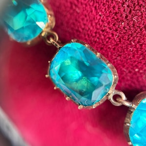 May include: A bracelet featuring a series of rectangular, faceted, bright turquoise-colored stones set in gold-toned metal frames. Each stone is connected by small metal links, creating a flexible chain design with a polished, reflective finish.