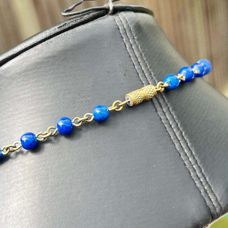 May include: A close-up of a necklace featuring vibrant blue beads strung on a delicate gold-toned chain. The necklace is fastened with a cylindrical clasp. The jewelry is displayed on a black surface.