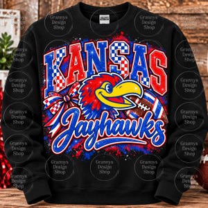 Pode incluir: Uma camisola preta de gola redonda com uma impressão gráfica com o texto KANSAS JAYHAWKS em vermelho, branco e azul. O design inclui uma cabeça de mascote de pássaro estilizada, uma bola de futebol americano e um laço xadrez sobre um fundo de salpicos.