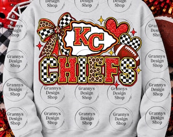 May include: A light grey crewneck sweatshirt featuring a graphic design with the text KC CHIEFS in red glitter and black and white chequered patterns. The design includes a large bow, a football, and heart shapes with leopard print accents.