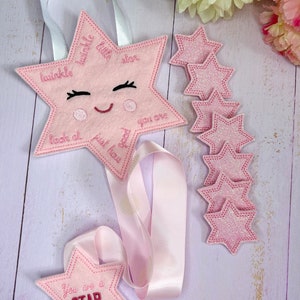 May include: Pink felt star with embroidered text "twinkle little star" and a smiling face. The star is attached to a pink ribbon and comes with six smaller pink felt stars with glitter.