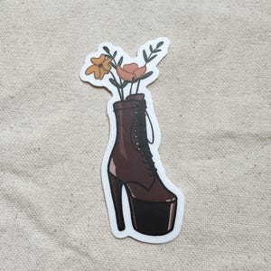 Könnte beinhalten: Ein Aufkleber mit einem braunen High-Heel-Stiefel mit Plateausohle, gefüllt mit orangefarbenen und gelben Blumen. Der Stiefel hat ein Schnürdesign und einen hohen, dünnen Absatz. Der Aufkleber befindet sich auf einem neutralfarbenen Stoffhintergrund.
