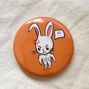 May include: Orange round button with a cartoon illustration of an angry white rabbit with a muscular physique. The rabbit has black eyes, whiskers, and a red mark on its forehead. A speech bubble with exclamation points is next to the rabbit.