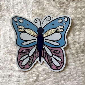 May include: A blue and pink butterfly sticker with glitter accents. The butterfly has a white body and black outlines.