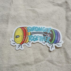 May include: A rainbow-colored barbell with the words "Stronger Together" written on it. The barbell is breaking through a cracked surface.
