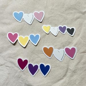 Op de afbeelding: Een verzameling hartvormige stickers in diverse kleuren, waaronder roze, blauw, geel, oranje, paars en zwart. Elke sticker toont drie verbonden harten met witte randen, geplaatst op een neutrale stoffen achtergrond.