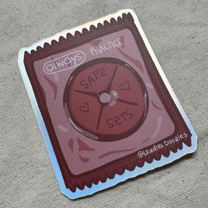 Könnte beinhalten: Ein Aufkleber mit Kondom-Design. Hauptsächlich rosa und rot, mit den Worten "always PRACTICE SAFE SETS" und einem kleinen Herzsymbol. Der Aufkleber hat einen holografischen Rand und den Text "@Leudles.Doodles".