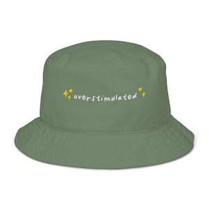 May include: Sage green bucket hat with the word "overstimulated" embroidered in white, accented by yellow starbursts. The hat has a wide brim and a simple, casual design.
