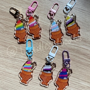 Könnte beinhalten: Sechs bunte Acryl-Schlüsselanhänger mit einem Comic-Charakter in einem Taiyaki mit Eiscreme. Jeder Anhänger hat ein anderes Farbschema und eine andere Flagge auf dem Eis. Die Anhänger sind an Metallschlüsselringen befestigt.