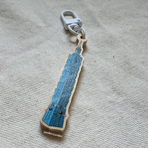 May include: A wooden keychain shaped like the Taipei 101 building with a blue and white colour scheme. The building has a cute cartoon face with black eyes and a pink blush. The keychain has a white metal clasp.