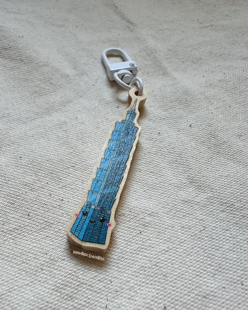 May include: A wooden keychain shaped like the Taipei 101 building with a blue and white colour scheme. The building has a cute cartoon face with black eyes and a pink blush. The keychain has a white metal clasp.
