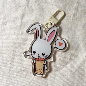 Pode incluir: Um chaveiro de acrílico transparente com um coelho de desenho animado bebendo bubble tea. O coelho é branco com orelhas rosa e um coração em um balão de fala. O chaveiro tem um fecho bege.