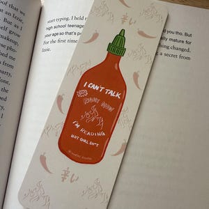 May include: A rectangular bookmark featuring a red sriracha bottle design with the text "I CAN'T TALK RIGHT NOW I'M READING HOT GIRL SH*T". The bookmark has a white background with chili pepper and flame illustrations.