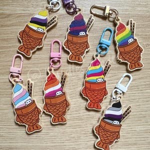 May include: Eight wooden keychain charms shaped like taiyaki, a Japanese fish-shaped pastry, with a cat peeking out of the top. Each taiyaki is filled with a different color of ice cream, representing different pride flags. The keychains are attached to silver, gold, and blue metal key rings.