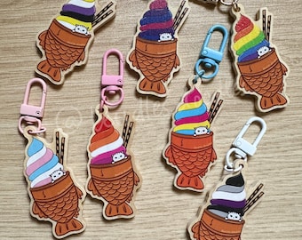 Puede incluir: Ocho llaveros de madera con forma de taiyaki, un pastel japonés con forma de pez, con un gato asomando por la parte superior. Cada taiyaki está relleno de un helado de diferente color, que representa diferentes banderas del orgullo. Los llaveros están unidos a llaveros metálicos plateados, dorados y azules.