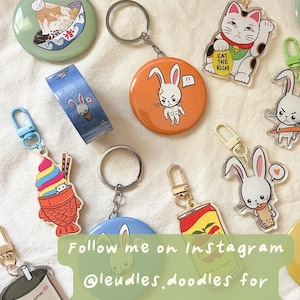 May include: Collection of colorful keychains and buttons with various designs, including a lucky cat, bunny characters, and a bubble tea cup. The image also includes a roll of washi tape with a bunny design. Text on the image reads "EAT THE RICH" and "EMOTIONAL SUPPORT".