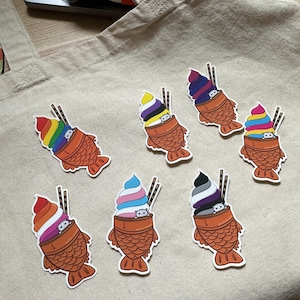 Pode incluir: Sete adesivos de cone de sorvete coloridos com um rosto de gato espiando pelo topo de cada cone. Os cones estão dentro de um cone de waffle em forma de peixe. Os adesivos estão em uma bolsa de lona bege.
