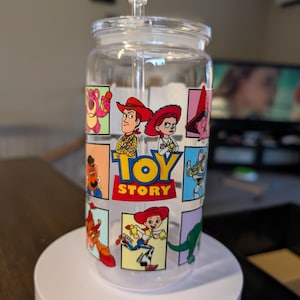 Puede incluir: Un vaso de cristal transparente con tapa y pajita que presenta una cuadr&iacute;cula colorida de personajes de Toy Story, incluidos Woody, Jessie y Buzz Lightyear. El gr&aacute;fico central muestra el logotipo de Toy Story en letras amarillas y rojas.