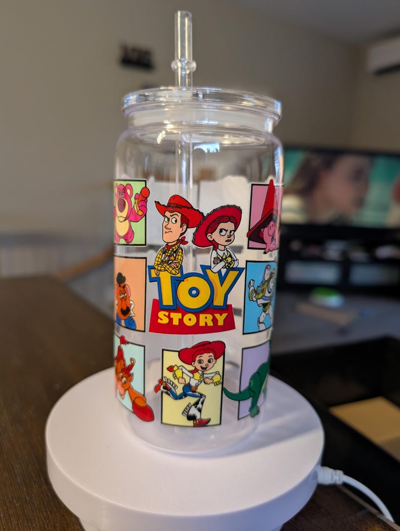 Puede incluir: Un vaso de cristal transparente con tapa y pajita que presenta una cuadr&iacute;cula colorida de personajes de Toy Story, incluidos Woody, Jessie y Buzz Lightyear. El gr&aacute;fico central muestra el logotipo de Toy Story en letras amarillas y rojas.