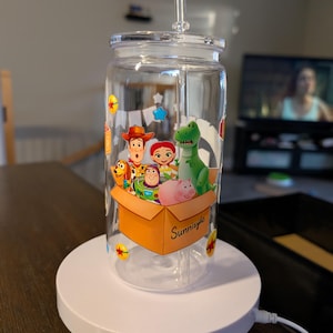 May include: A clear glass tumbler with a lid and straw featuring a colorful graphic of cartoon characters including a cowboy, space ranger, cowgirl, green dinosaur, and pink pig inside a cardboard box labeled Sunnyside with small star accents.